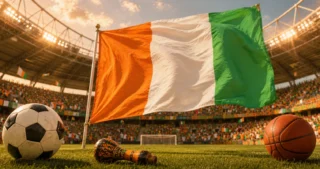 Drapeau de la Côte d’Ivoire flottant dans un stade, avec ballons de football et de basket au premier plan.